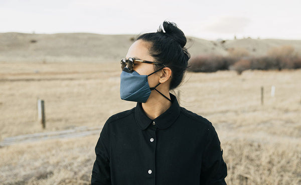 Person standing outdoors in a grassy field wearing the V6 Mask in Pacific Blue. The person is wearing sunglasses.